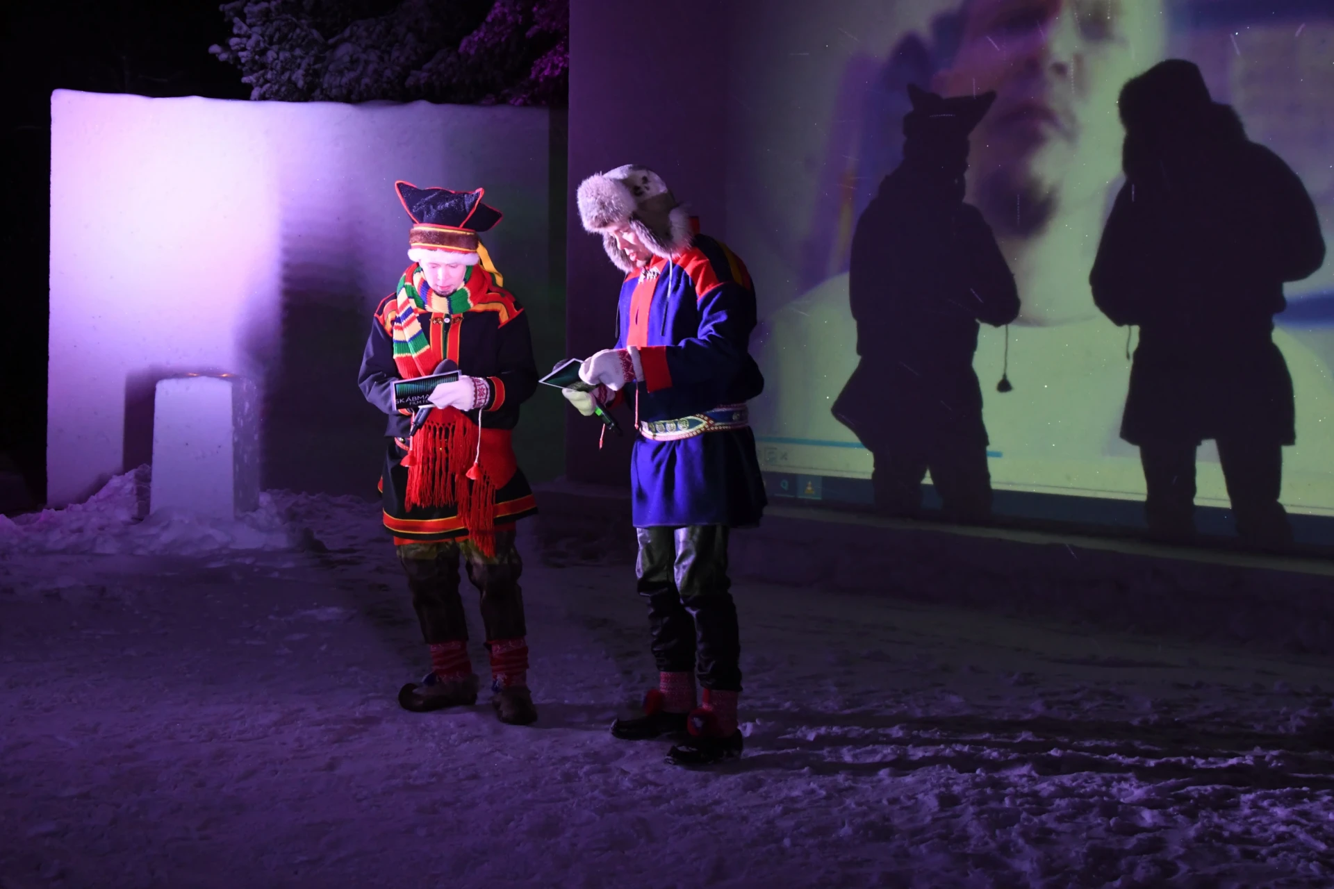 Two Sámi in front of a screen made of snow.